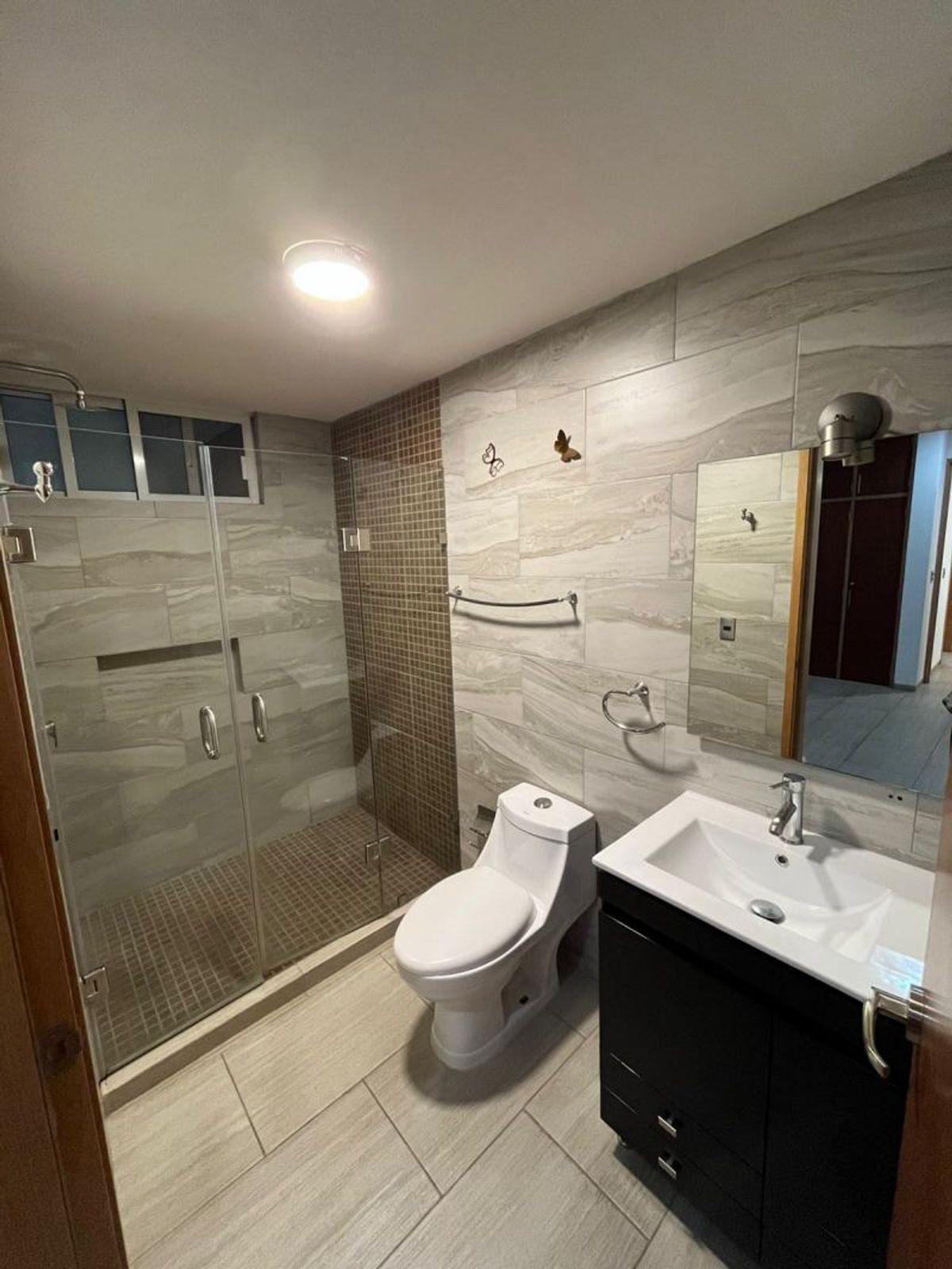 Baño con mueble de lavabo oscuro, inodoro blanco y regadera con cancel de cristal