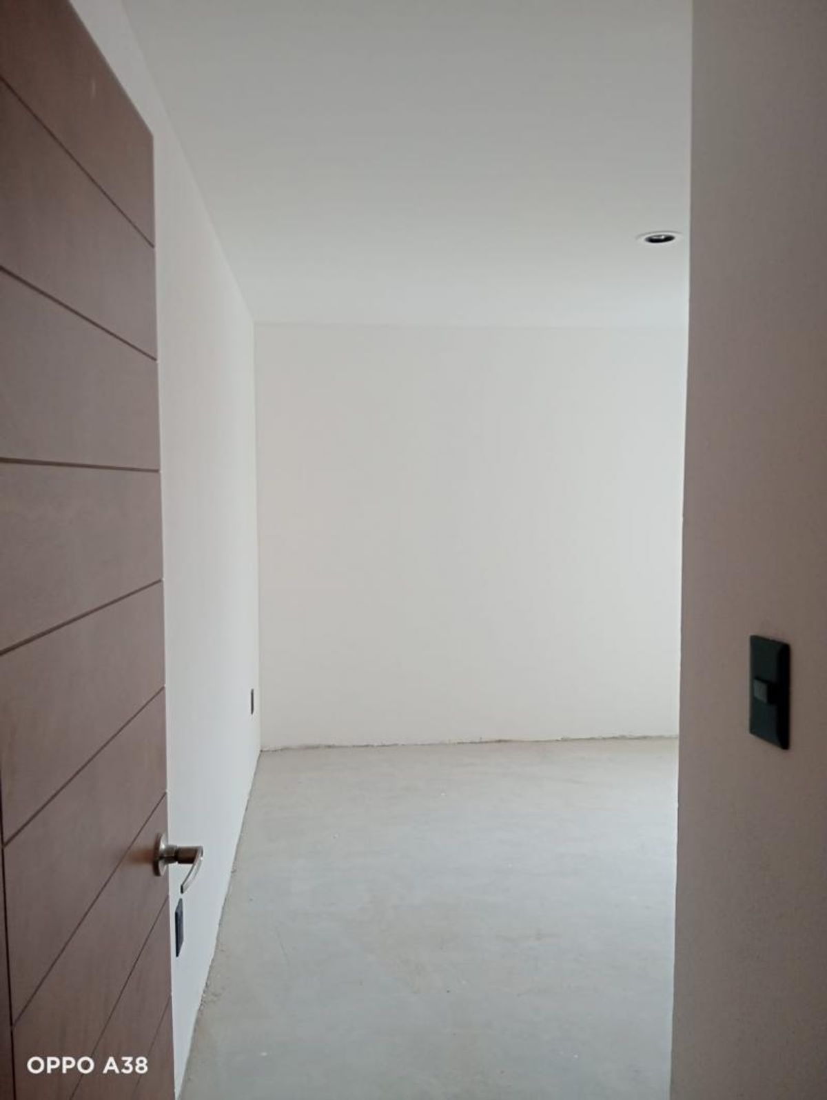 Habitación vacía con paredes blancas, piso gris claro, luz empotrada y puerta de madera