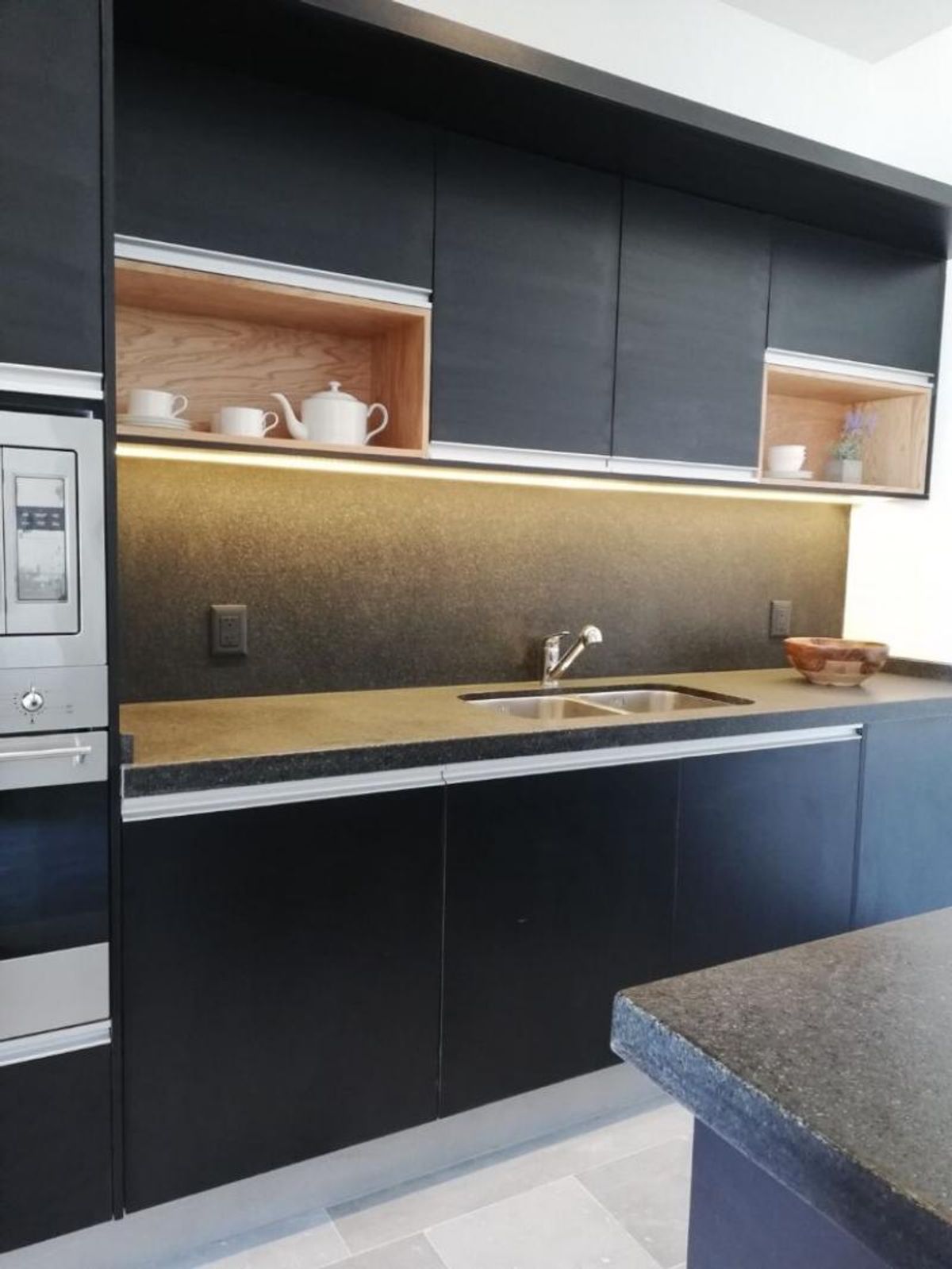 Cocina con gabinetes negros, encimera oscura, iluminación bajo alacenas y nichos de madera clara