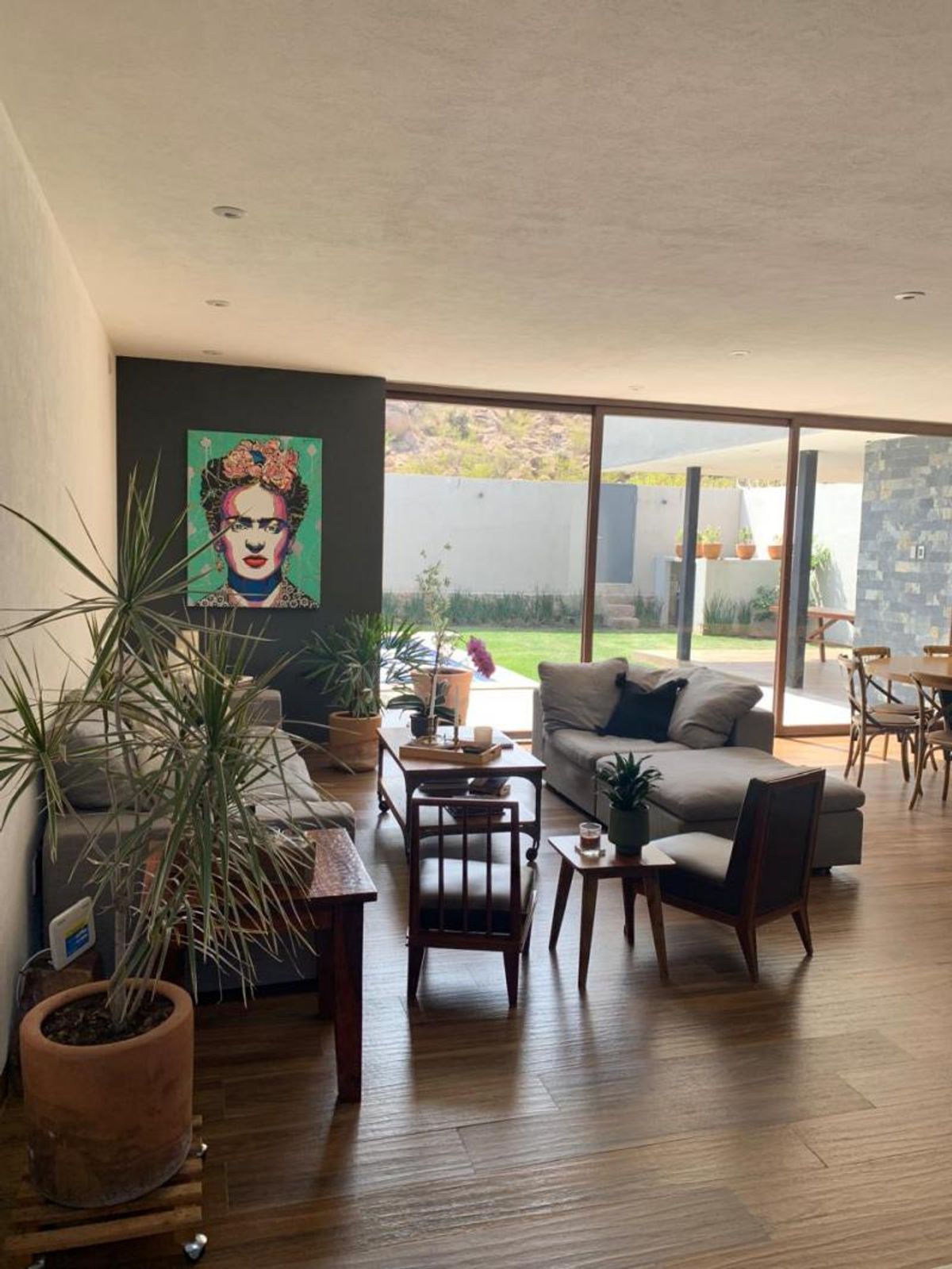 Sala con sofás grises, piso de madera, plantas decorativas y ventanales hacia el jardín