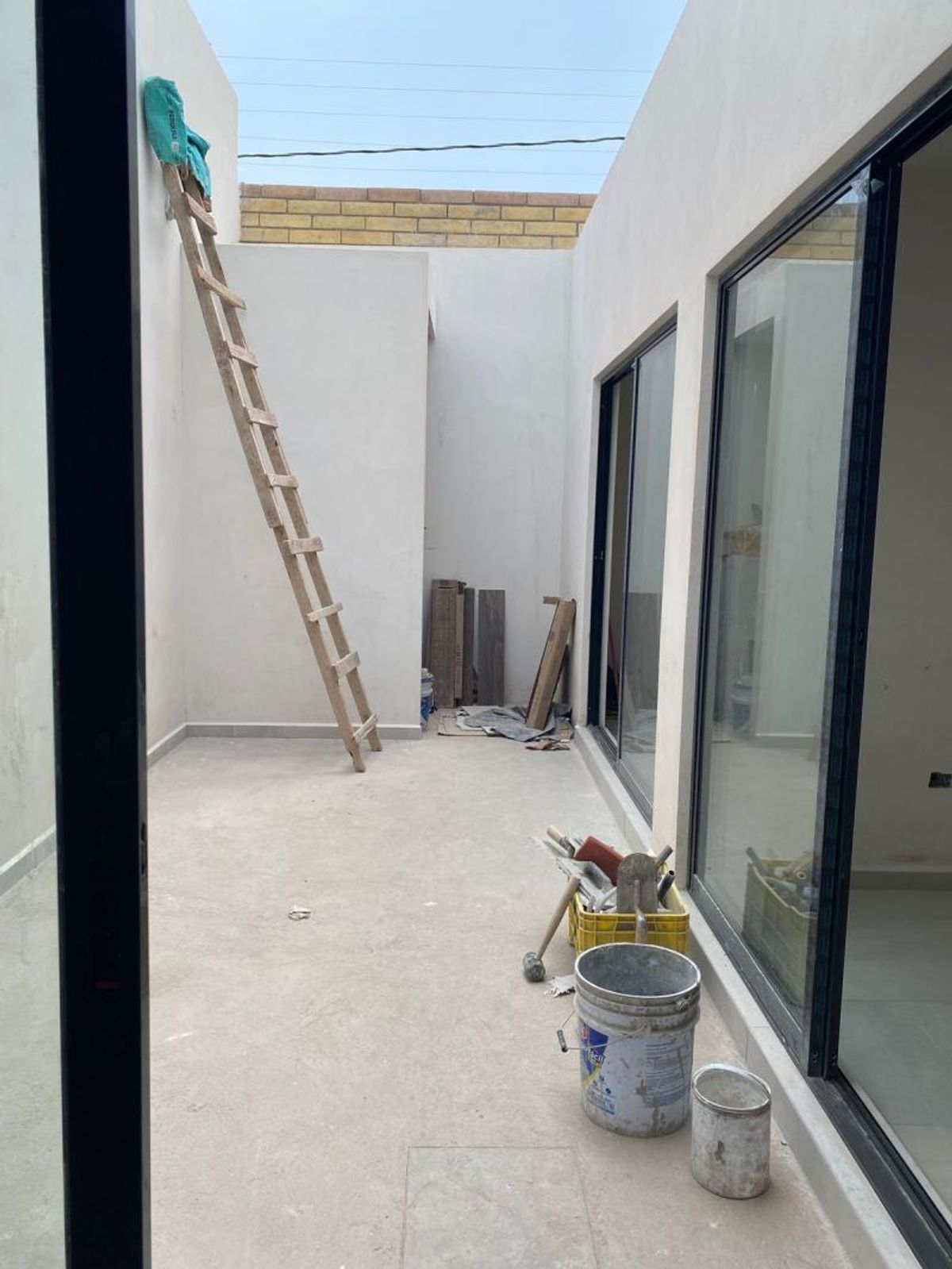 Patio interior en construcción con paredes blancas, ventanales corredizos negros y herramientas de obra