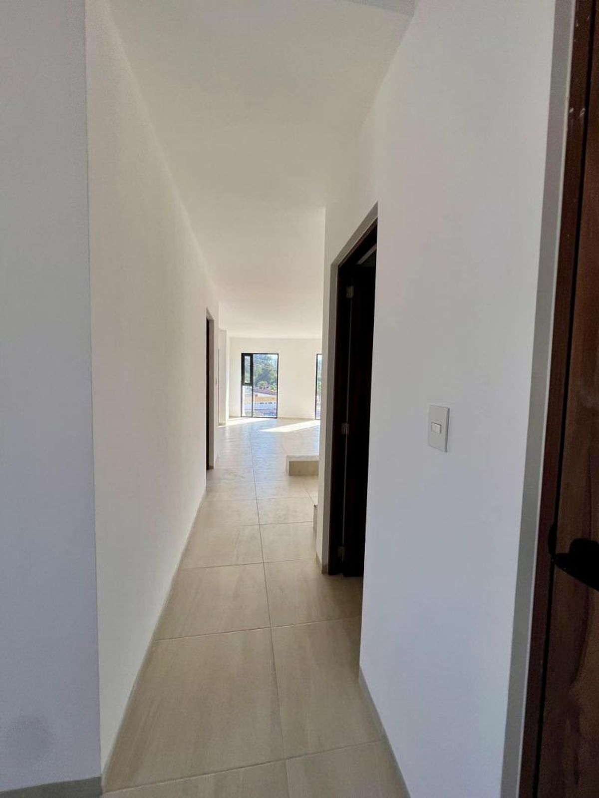 Pasillo blanco con piso cerámico claro, puertas de madera y ventana al fondo