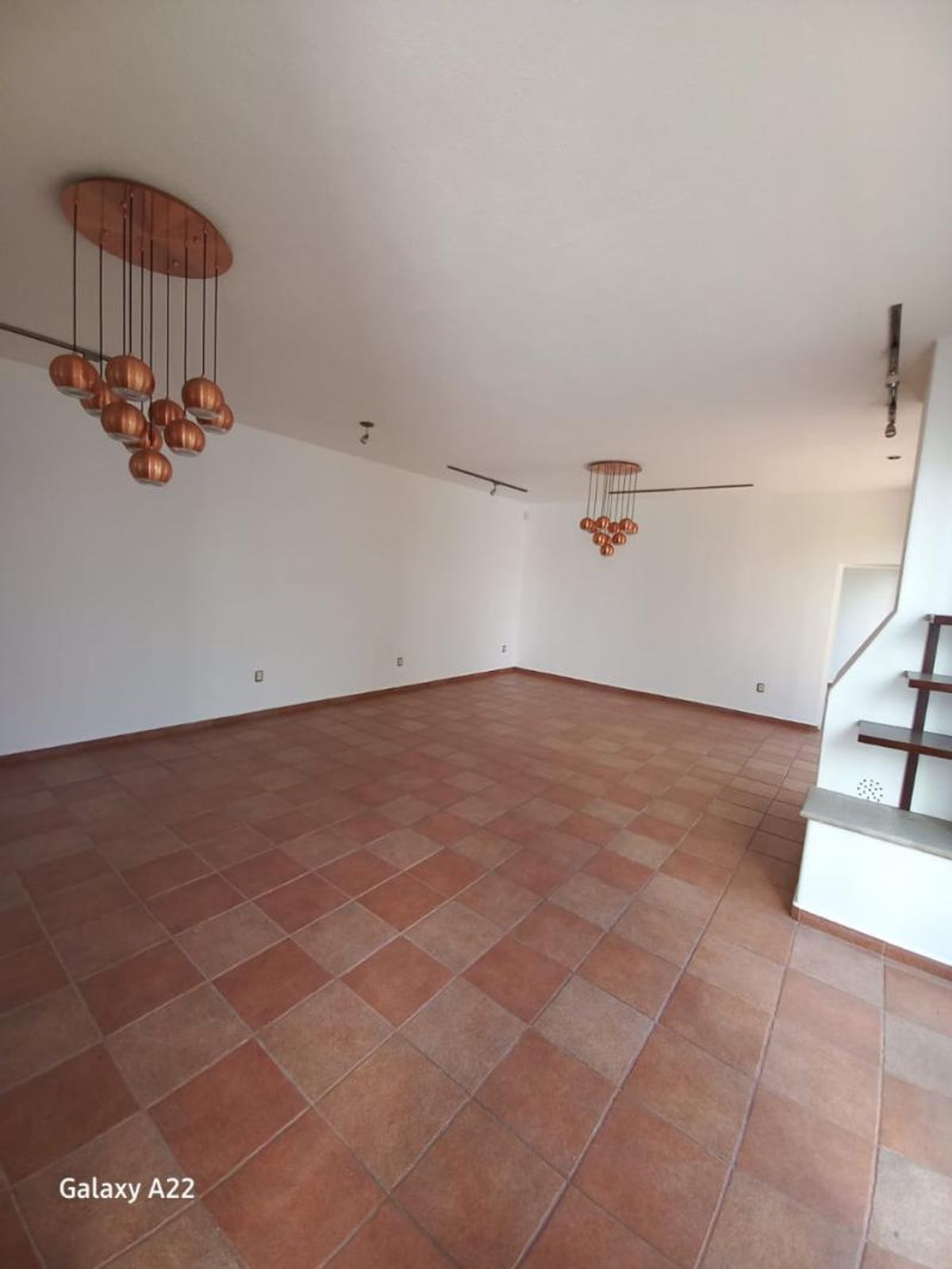 Área vacía con piso de loseta terracota, paredes blancas y lámparas colgantes de cobre