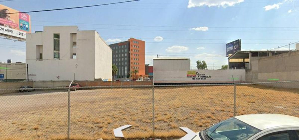 Terreno plano con pasto seco delimitado por malla ciclónica y edificios al fondo
