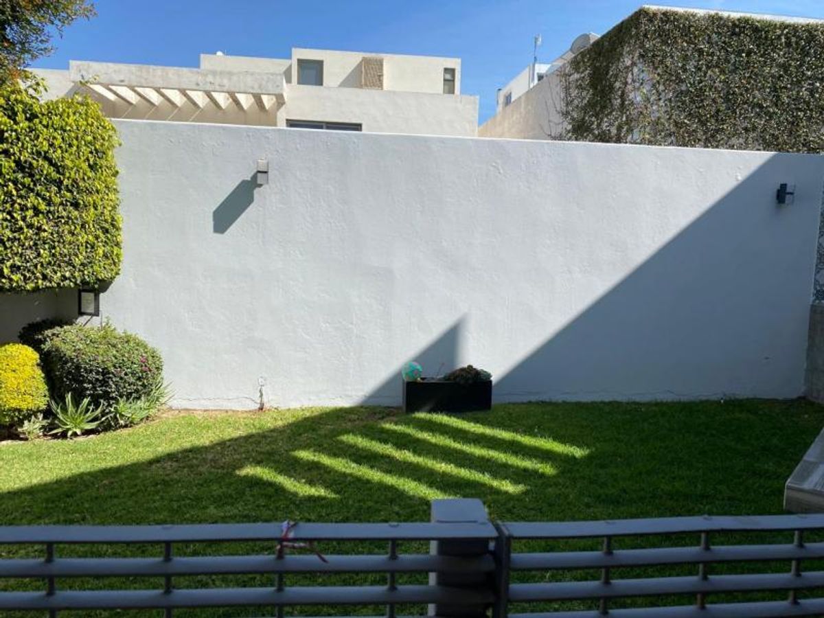 Jardín con césped verde, muro perimetral blanco, apliques de luz y arbustos laterales