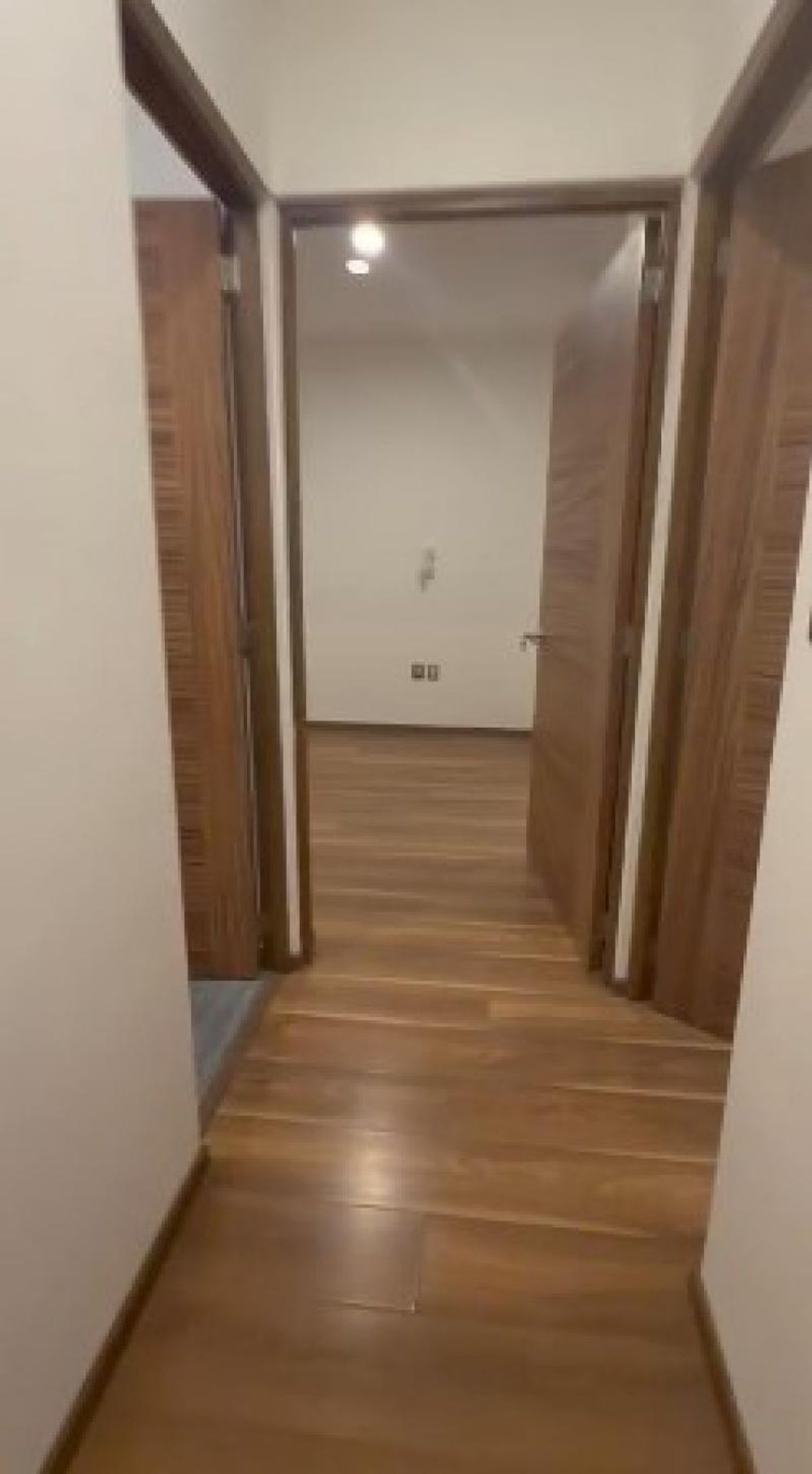 Pasillo con piso laminado de madera, paredes blancas y puertas abiertas hacia una habitación