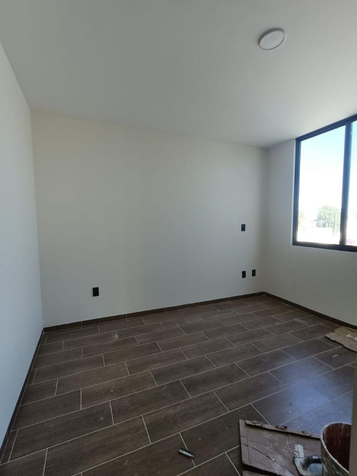 Habitación vacía con piso cerámico tipo madera, paredes claras, contactos negros y ventana lateral