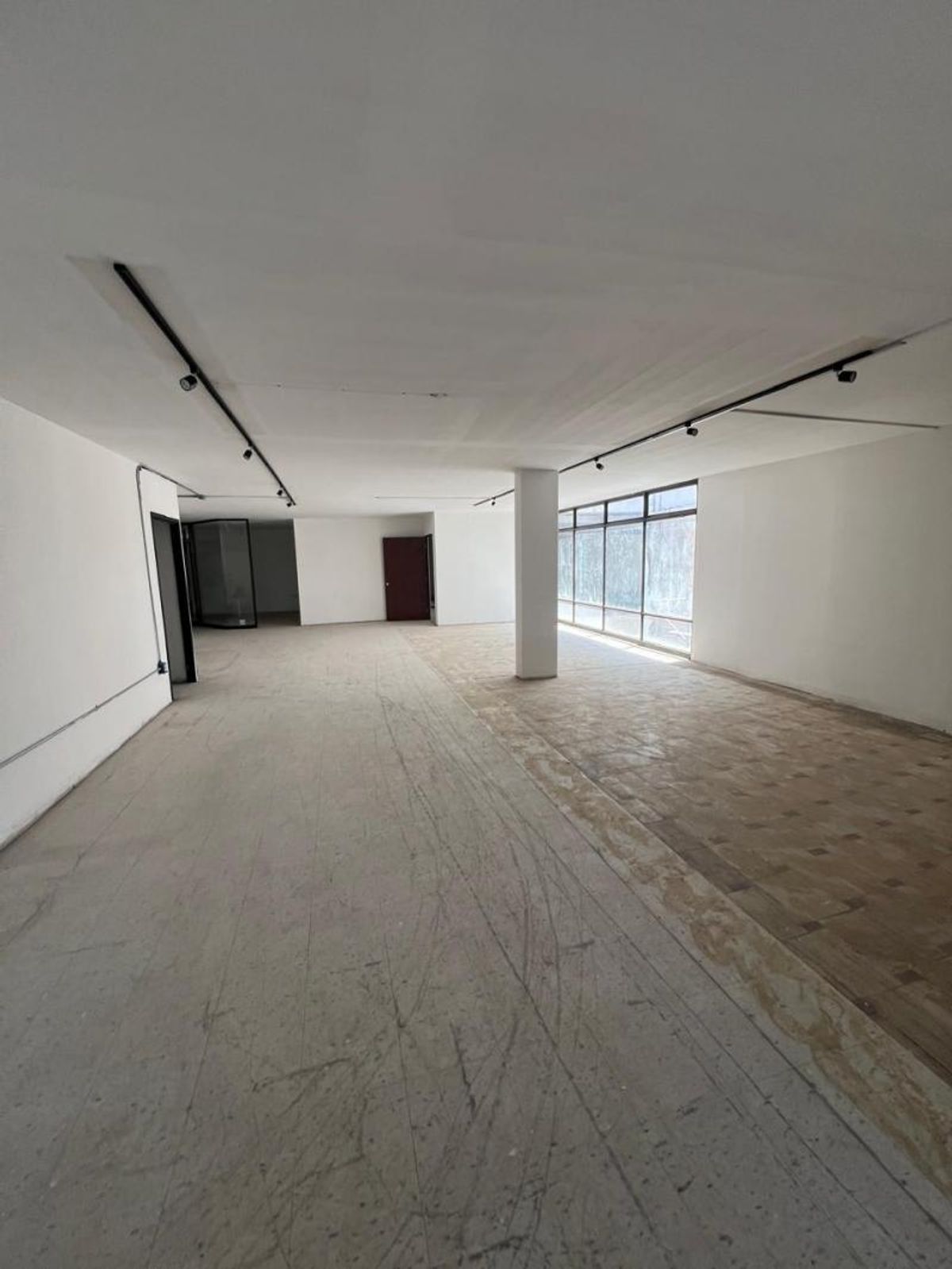 Espacio amplio y vacío con paredes blancas, columna central, rieles de iluminación y ventanales