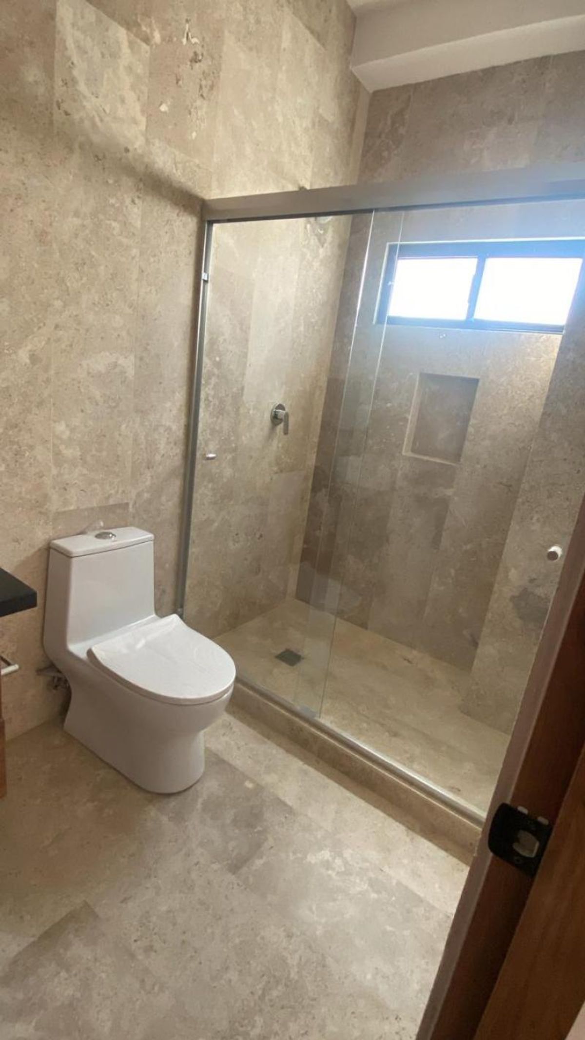 Baño revestido en piedra beige con inodoro blanco y regadera con cancel de cristal