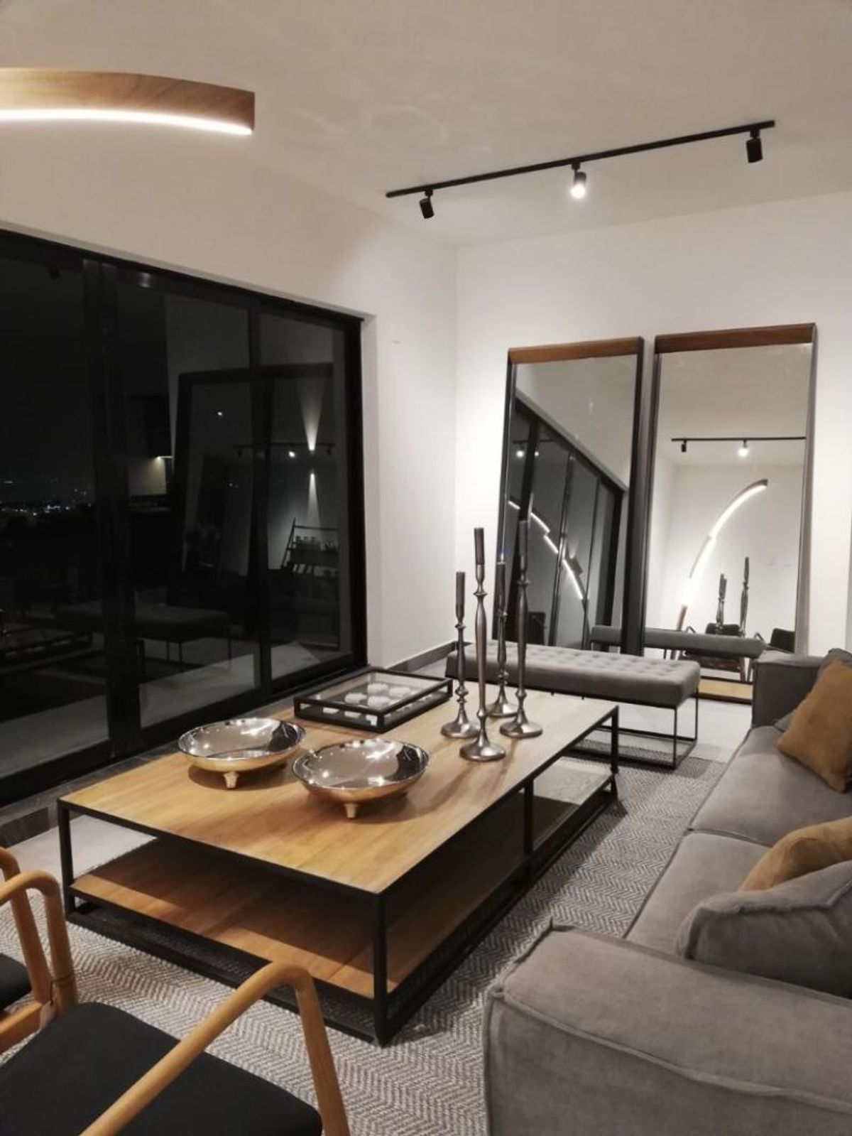 Sala con mesa de centro de madera, sofá gris, espejos de pie y ventanales