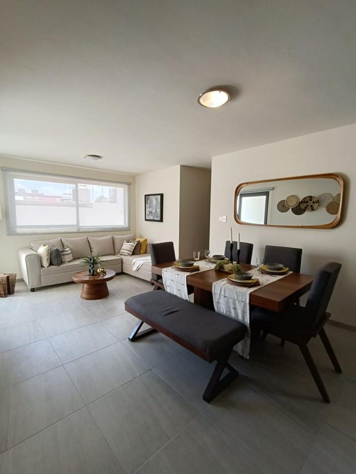 Comedor con mesa de madera, banca tapizada, sala con sofá modular y piso cerámico claro