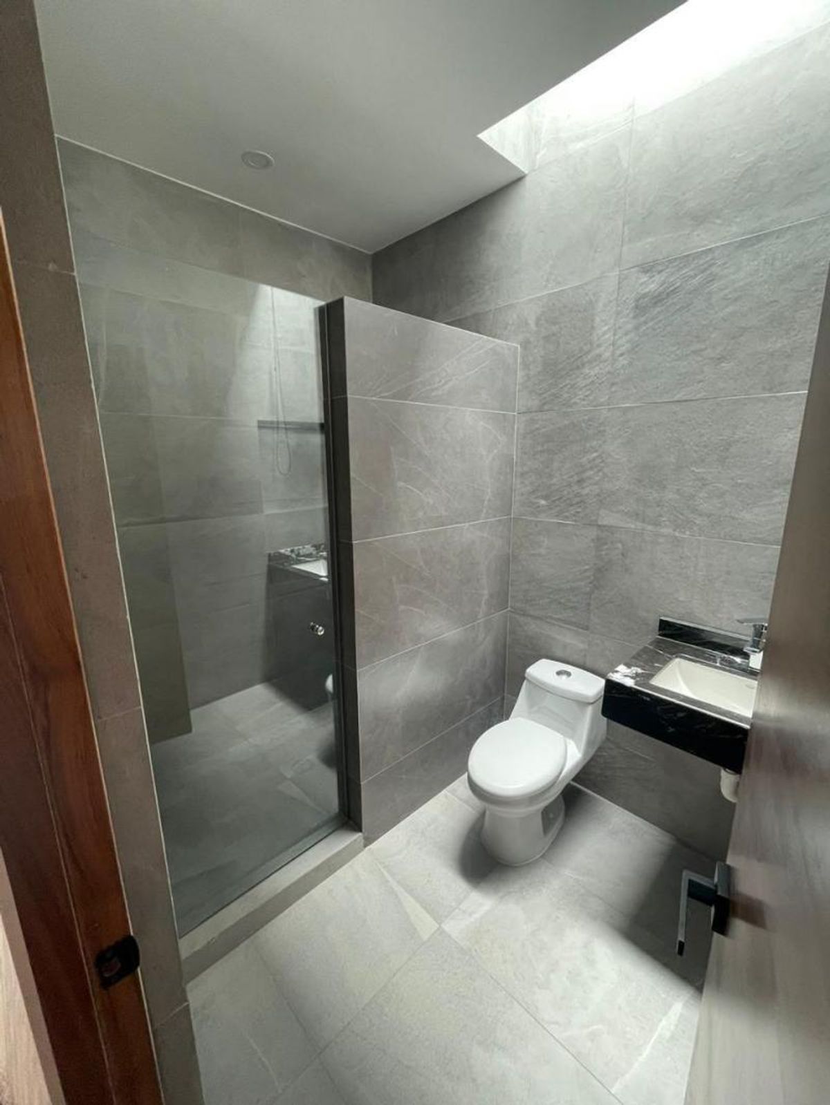 Baño con paredes de azulejo gris, regadera con cristal, inodoro blanco y tragaluz superior
