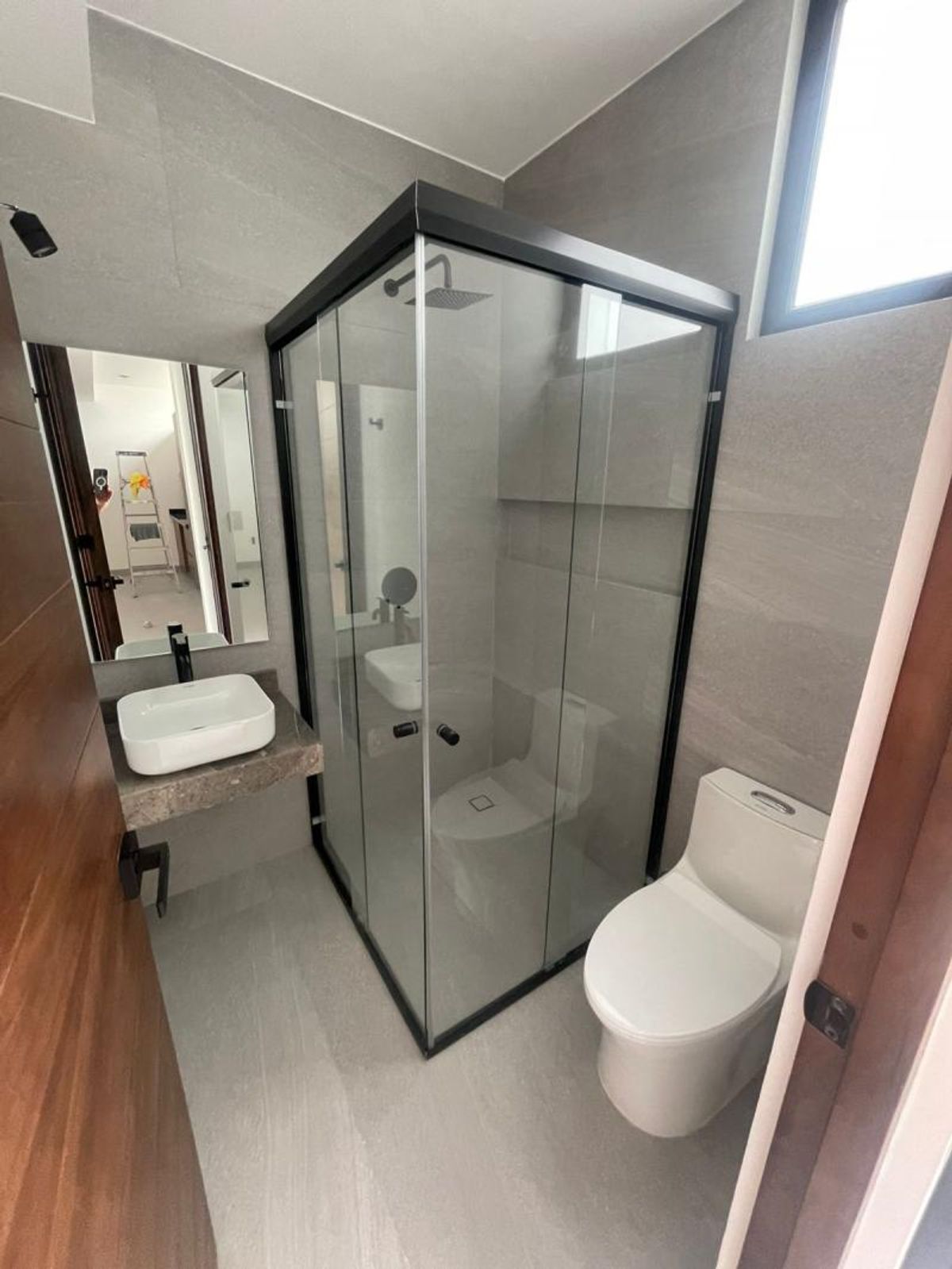 Baño con revestimiento gris, cancel de cristal negro, inodoro y lavabo de sobreponer
