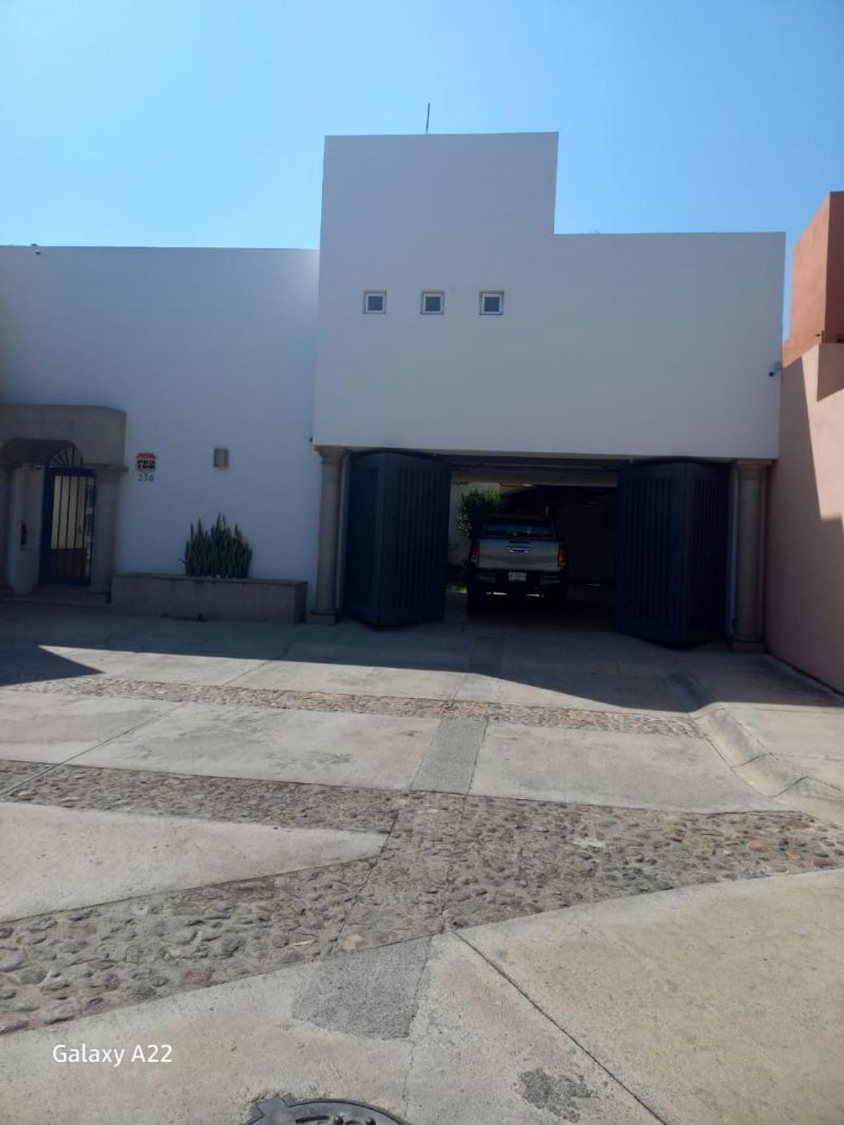 Fachada blanca con cochera abierta, portón oscuro y piso exterior de concreto con empedrado