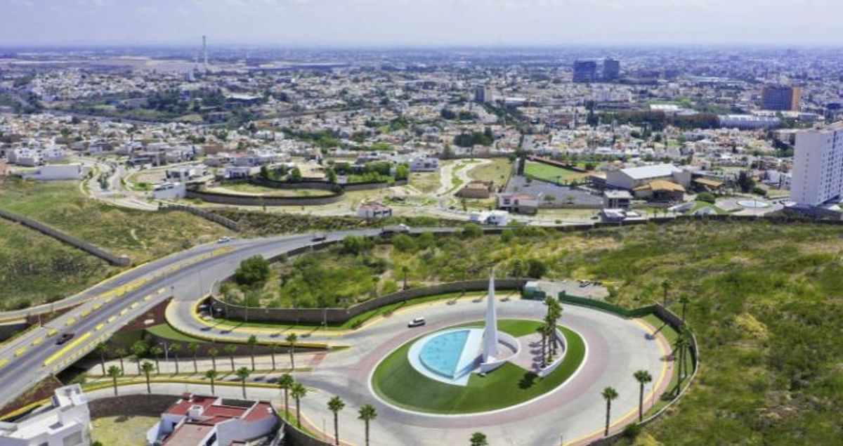 Glorieta circular con monumento blanco, fuente azul, palmeras y vialidades de un fraccionamiento residencial