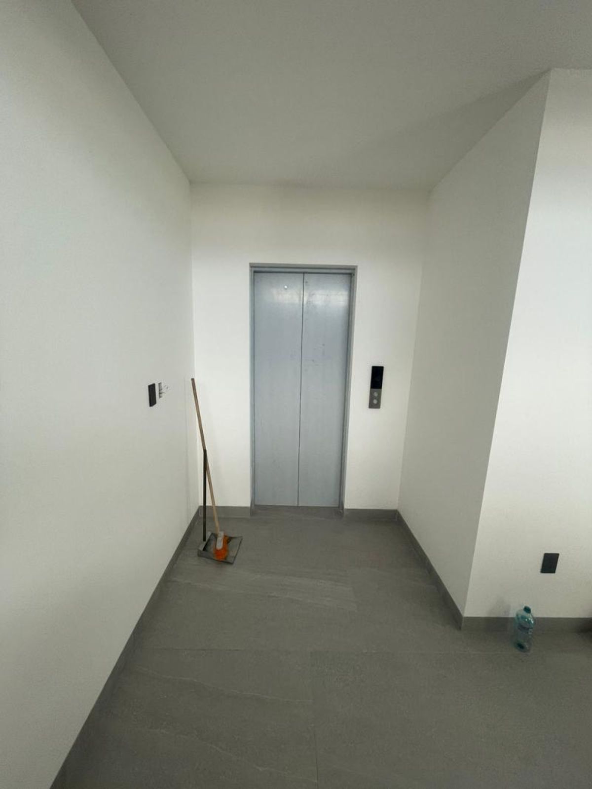 Pasillo con elevador de puertas metálicas, paredes blancas y piso cerámico gris