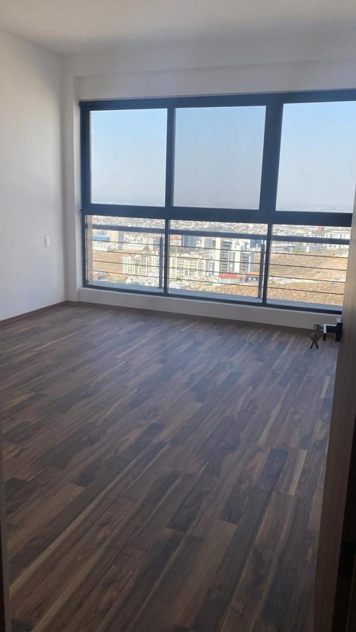 Habitación vacía con piso laminado de madera, paredes blancas y ventanal con vista urbana