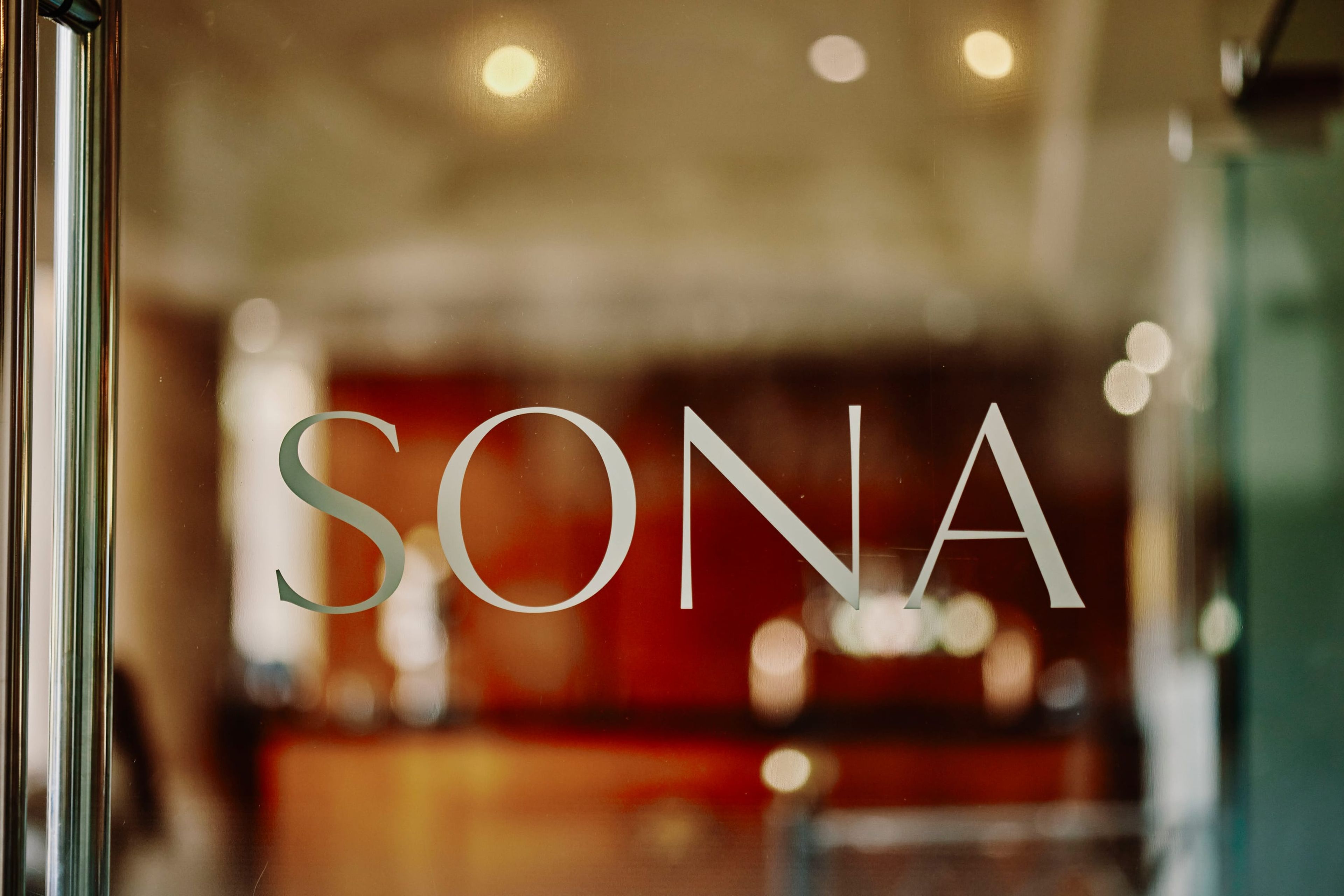 The SONA name etched on the glass entrance, with the warm dining room glowing beyond