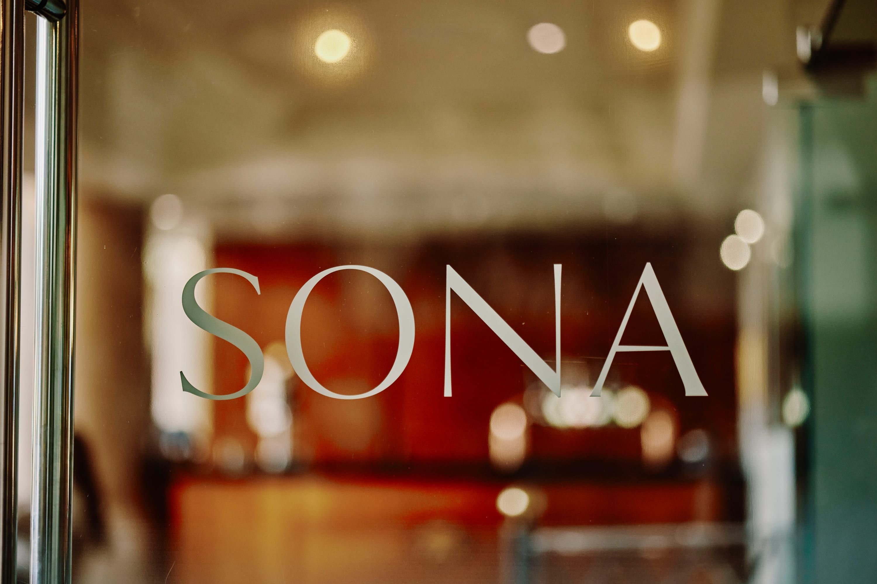 The SONA name etched on the glass entrance, with the warm dining room glowing beyond