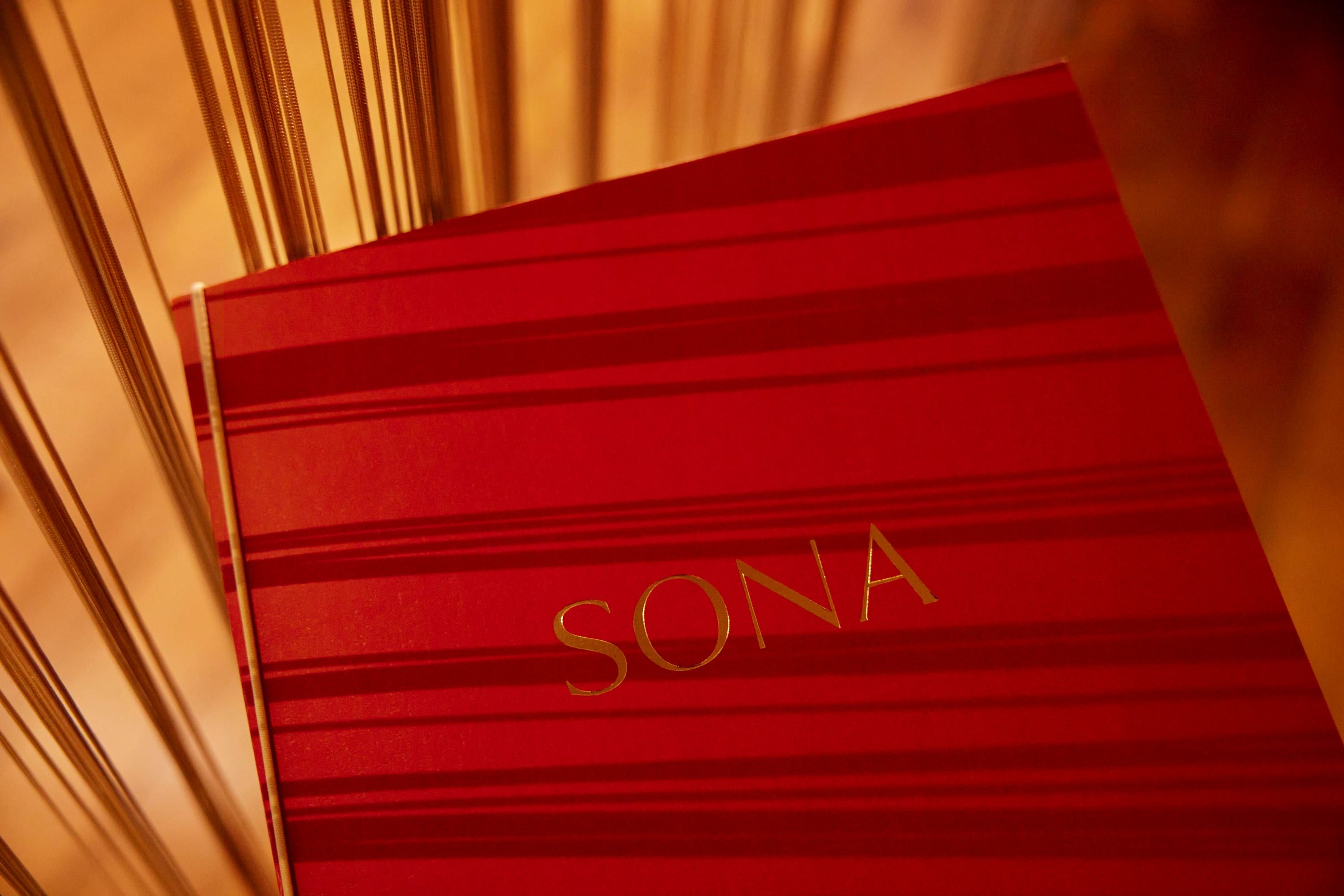 The red SONA menu book with gold lettering, lit by warm light