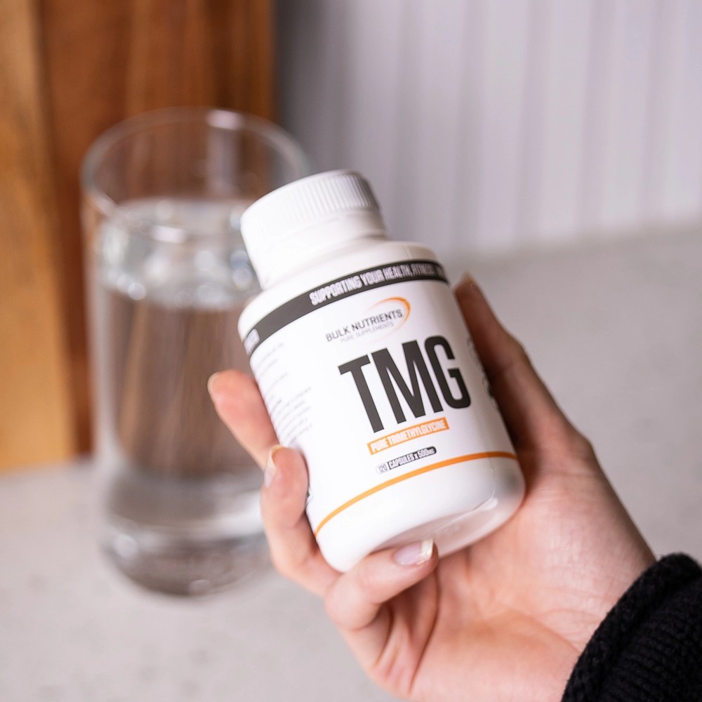 Bulk Nutrients TMG Capsules being held in front of a glass of water