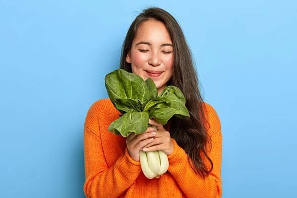 A women wearing a bright orange sweater smiling and holding a fresh looking bunch of bok choy.