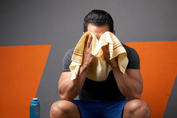 A muscular man resting between sets on a gym bench while wiping the sweat off his face with his towel. 