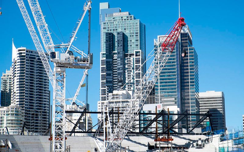 Construction cranes towering over a Sydney CBD building site, with high-rise office and residential towers in the background under a clear blue sky.