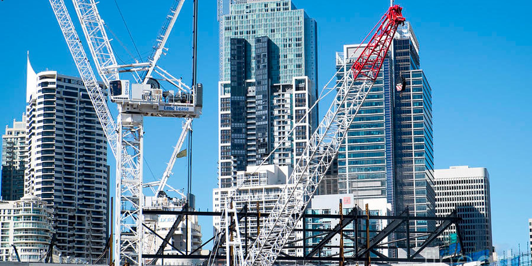 Construction cranes towering over a Sydney CBD building site, with high-rise office and residential towers in the background under a clear blue sky.