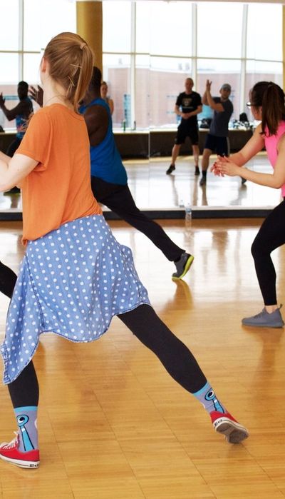 Tanzunterricht wird von hinten gezeigt in einem Tanzstudio. Vorne links tanz ein Mädchen mit orangenem T-shirt und blau- weiß gepunkteter Jacke um die Hüfte. 