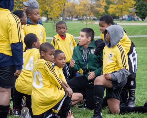 Un entraîneur en veste verte s'accroupit devant son équipe de jeunes footballeurs. Les enfants portent tous des maillots jaunes.