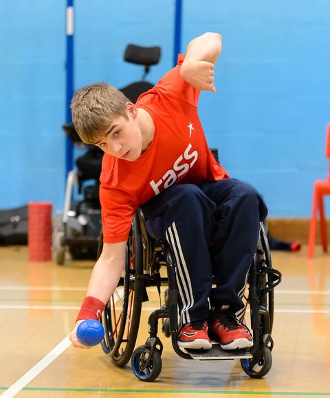 Un garçon en fauteuil roulant portant un T-shirt orange et un long pantalon de sport bleu. Il se penche sur le côté pour ramasser une petite balle bleue.