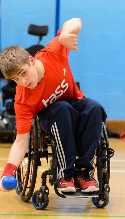 Ein Junge in einem Rollstuhl mit orangenem T-Shirt und blauer, langer Sporthose. Er bückt sich mit dem Oberkörper zur Seite um einen kleinen blauen Ball aufzuheben.