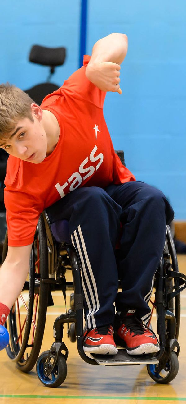 Ein Junge in einem Rollstuhl mit orangenem T-Shirt und blauer, langer Sporthose. Er bückt sich mit dem Oberkörper zur Seite um einen kleinen blauen Ball aufzuheben.