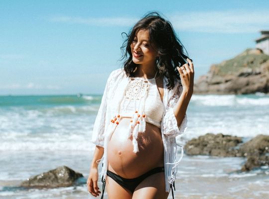 a pregnant lady walking along a sunny beach playing with her hair 