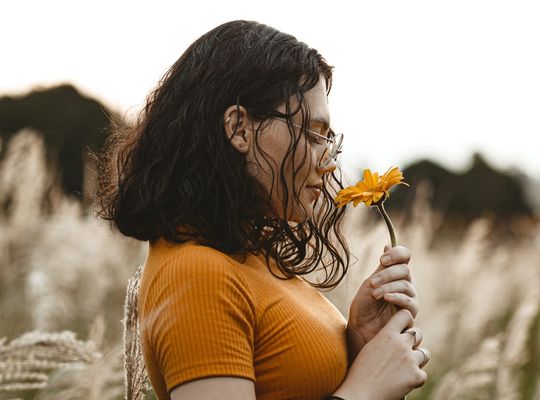 A girl in a yellow top standing in a field smelling a yellow flower 