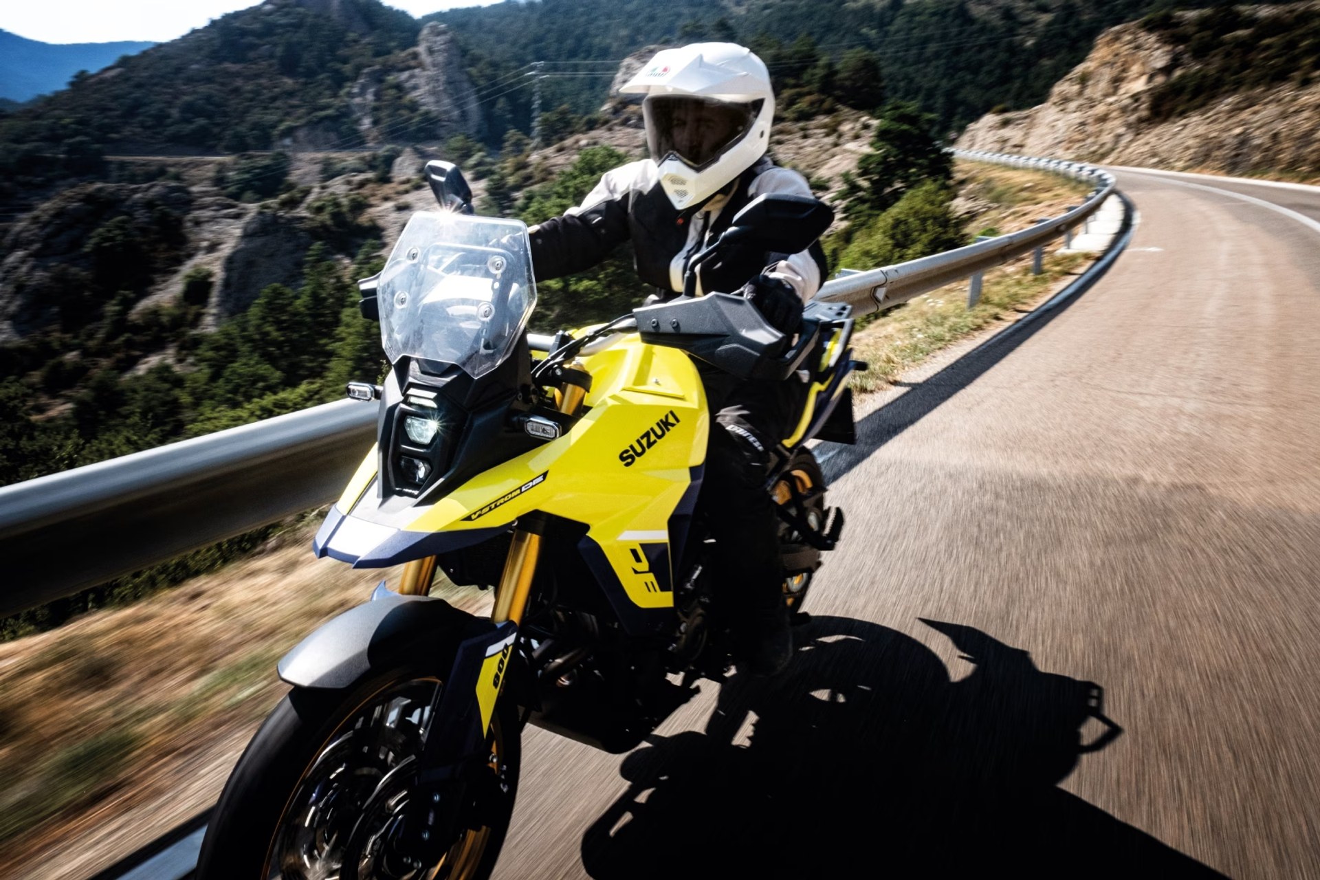 Piloto con casco blanco conduciendo una moto Suzuki V-Strom 800DE amarilla en carretera de montaña