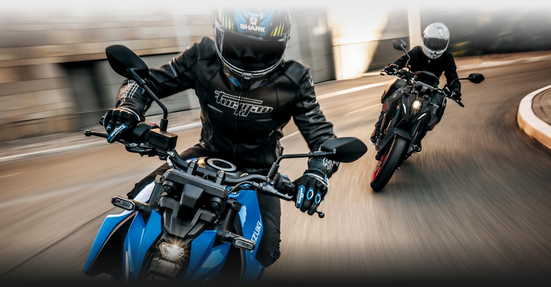 Dos motociclistas conduciendo motos Suzuki deportivas por una carretera urbana
