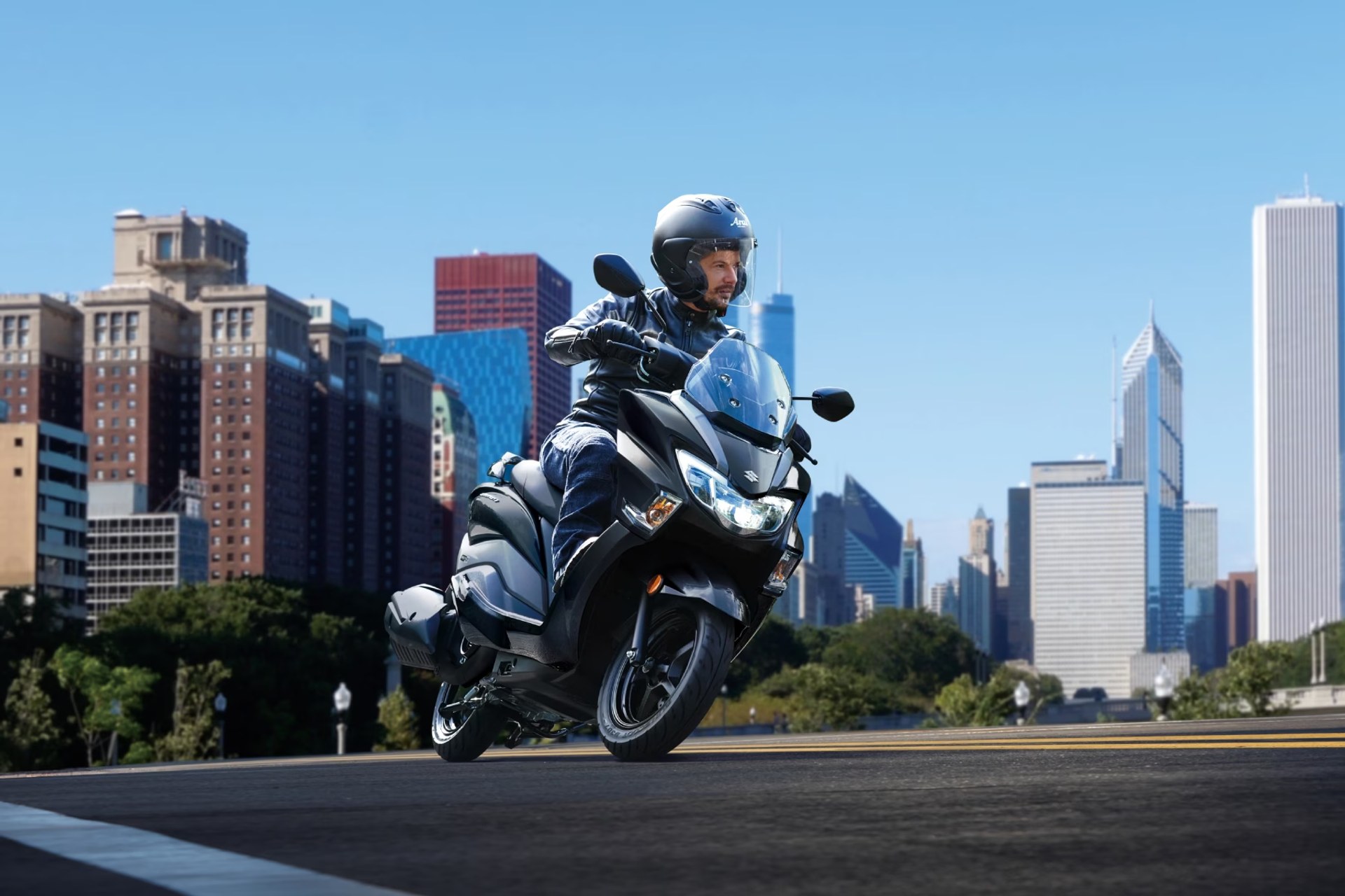 Piloto conduciendo un scooter Suzuki Burgman Street 125EX por la ciudad con rascacielos de fondo