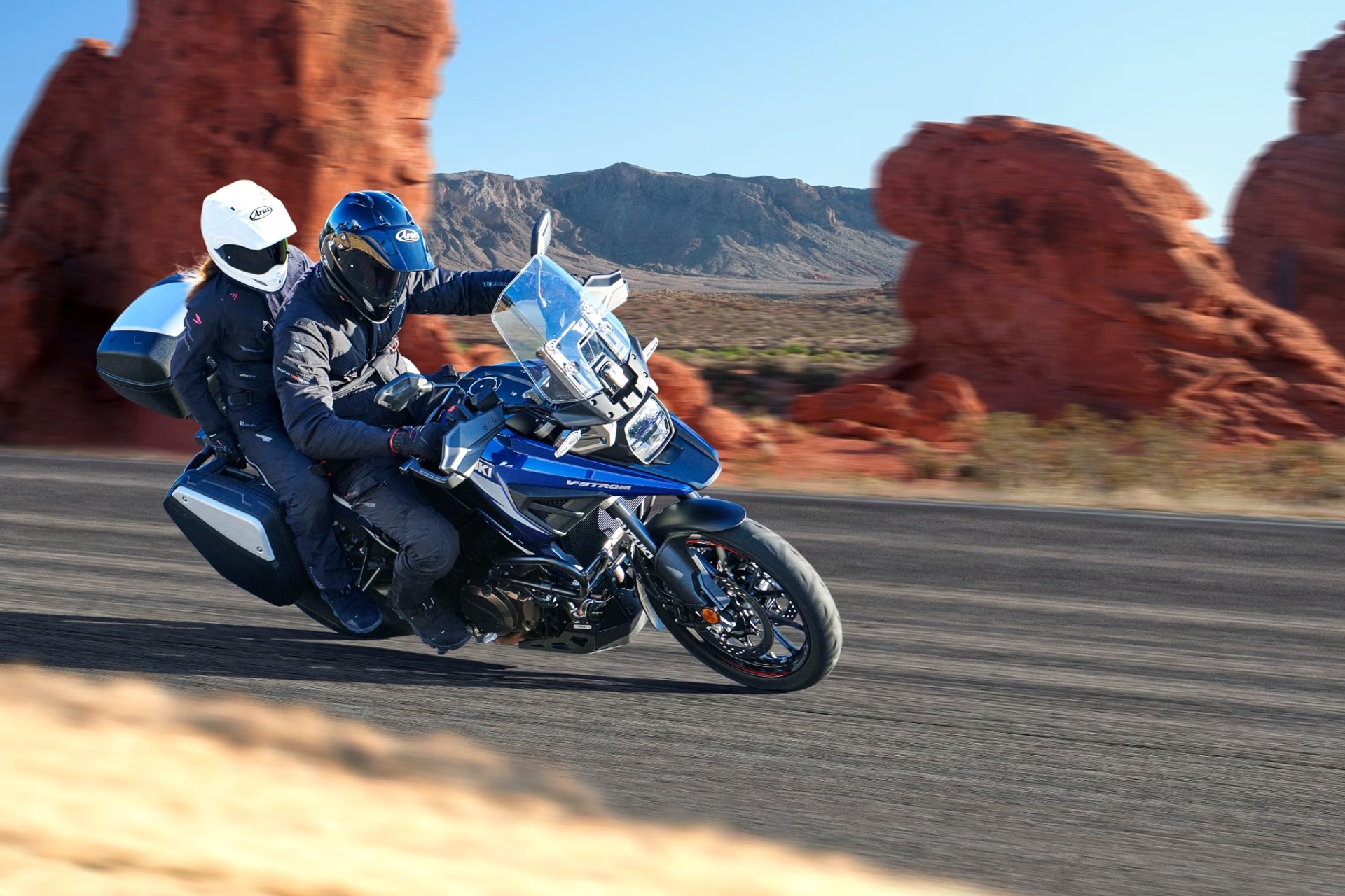 Dos motociclistas en Suzuki V-Strom 1050DE azul recorriendo una carretera entre formaciones rocosas