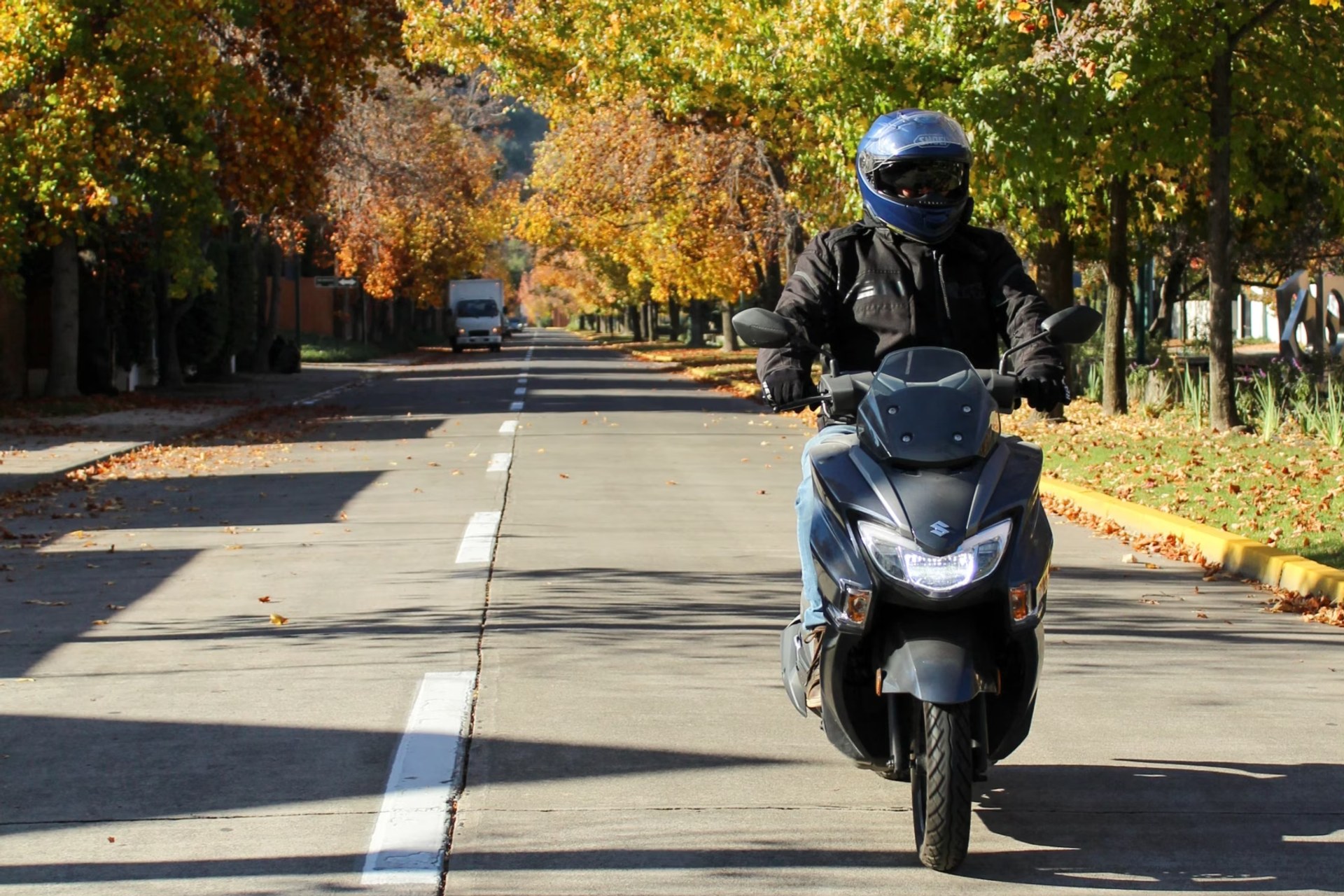 Piloto conduciendo un scooter Suzuki Burgman Street 125EX gris por el centro de una carretera rodeada de árboles