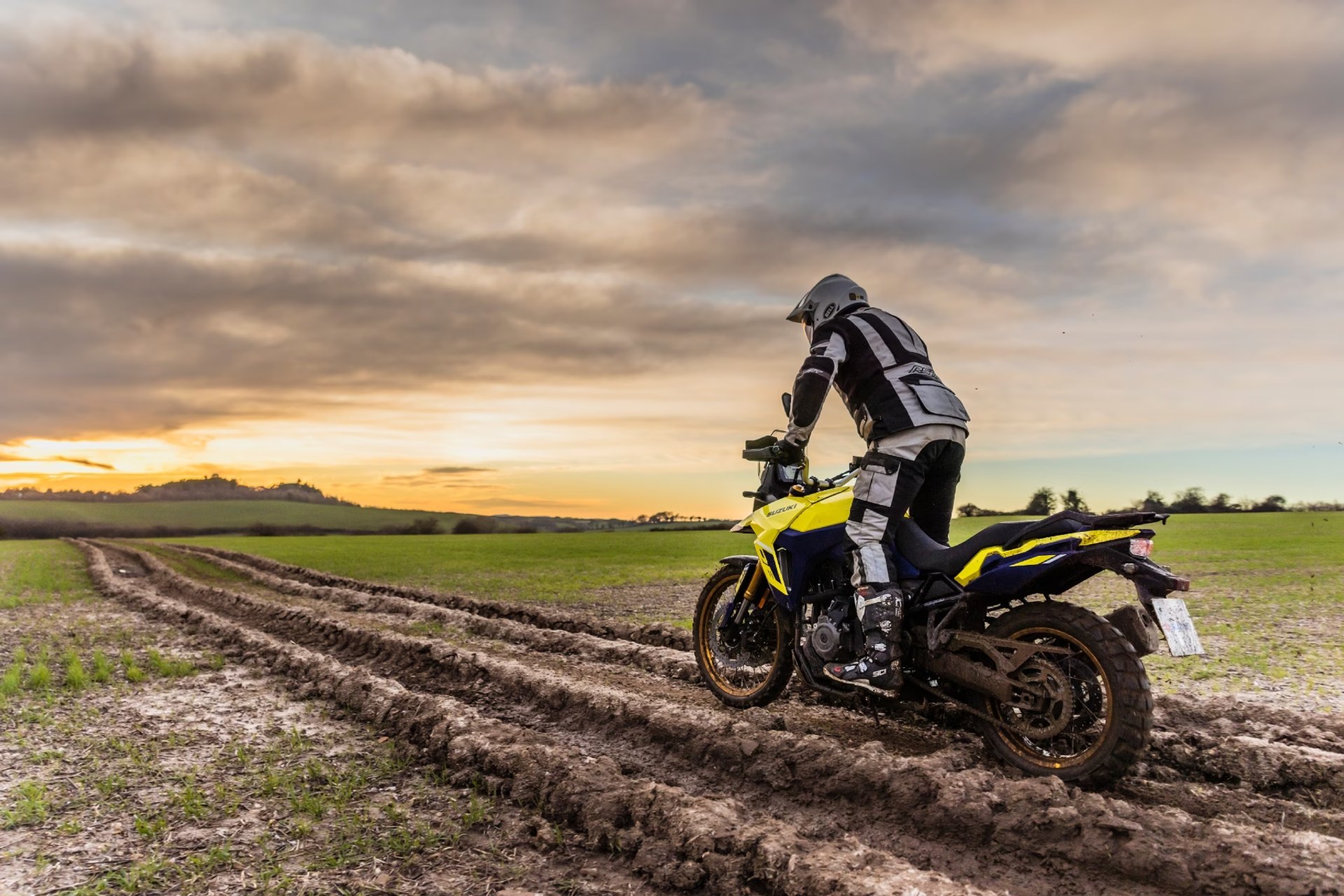 Piloto con equipación completa conduciendo una moto de aventura Suzuki V-Strom 800DE por un camino de barro profundo en el campo al atardecer