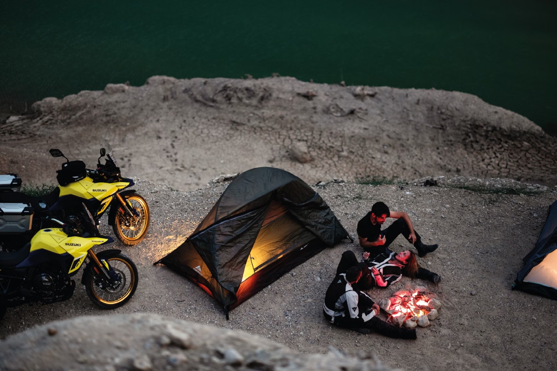 Motociclistas acampando junto a sus motos Suzuki V-Strom 800DE alrededor de una fogata al anochecer