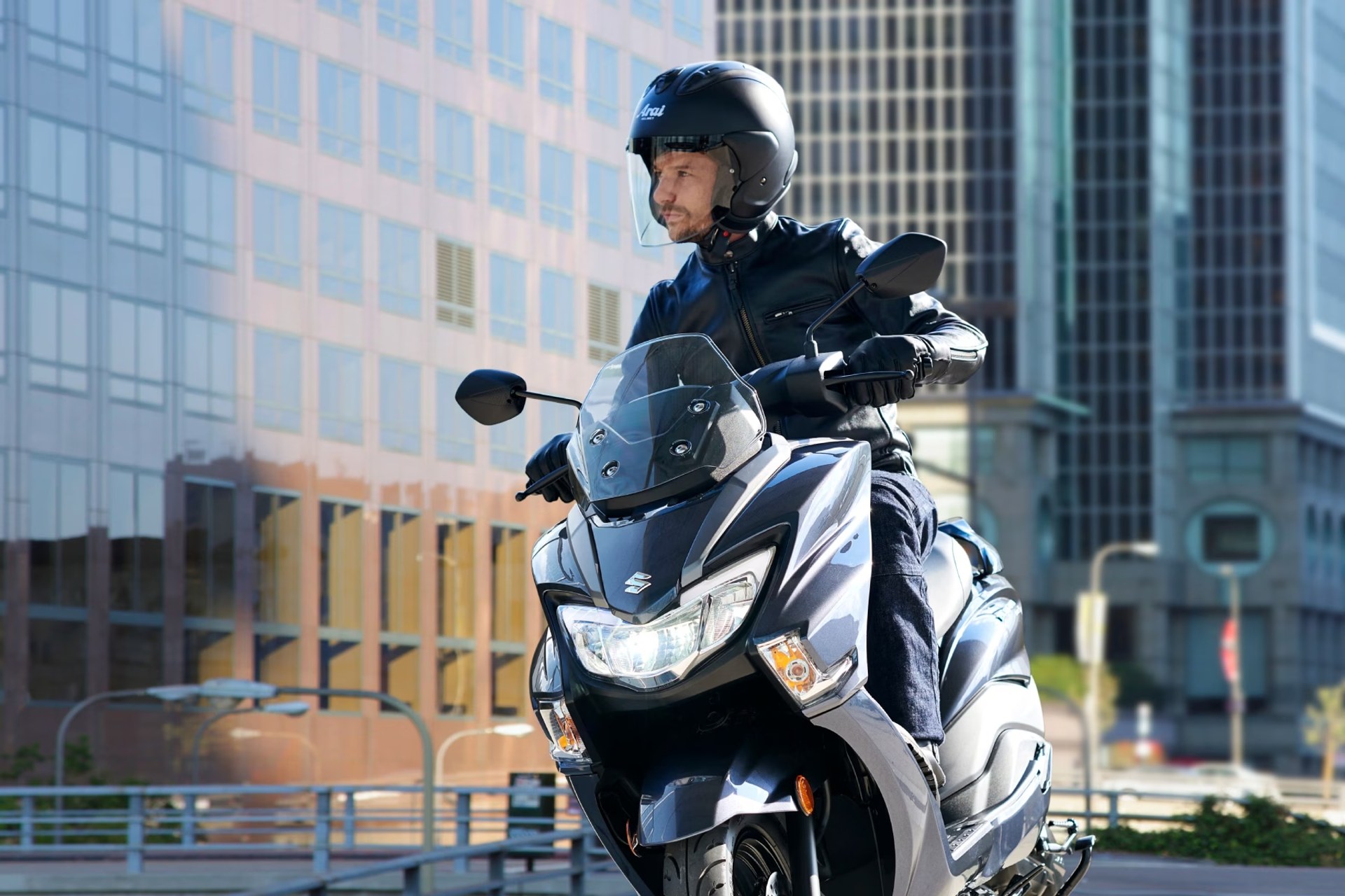 Piloto conduciendo un scooter Suzuki Burgman Street 125EX gris por la ciudad entre edificios