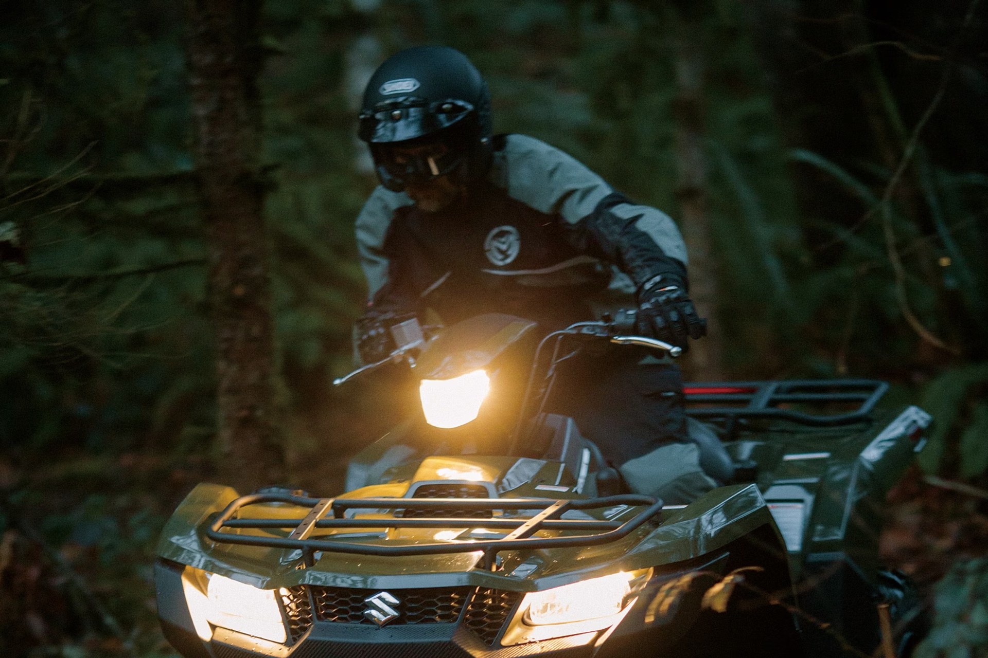 Conductor en Suzuki KingQuad 750 verde recorriendo un sendero de bosque con luces encendidas