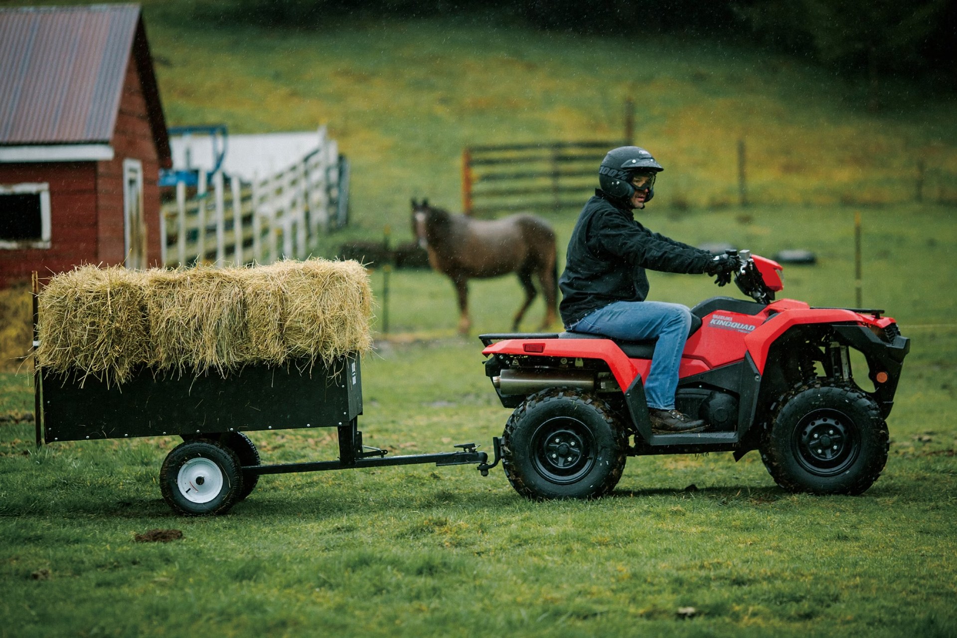 Conductor en Suzuki KingQuad 750 rojo remolcando fardos de heno en un entorno rural