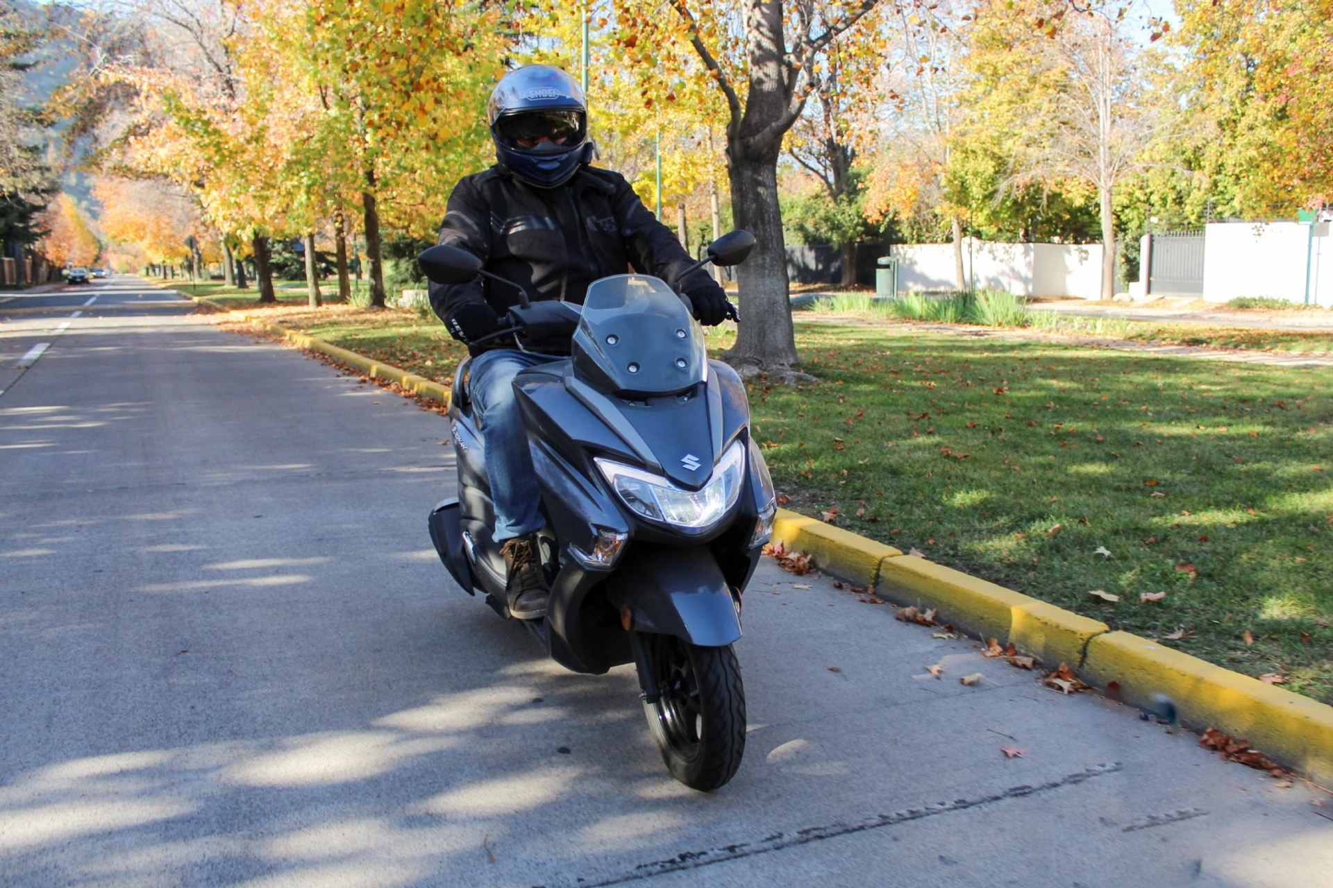 Piloto conduciendo un scooter Suzuki Burgman Street 125EX gris por una avenida residencial en otoño