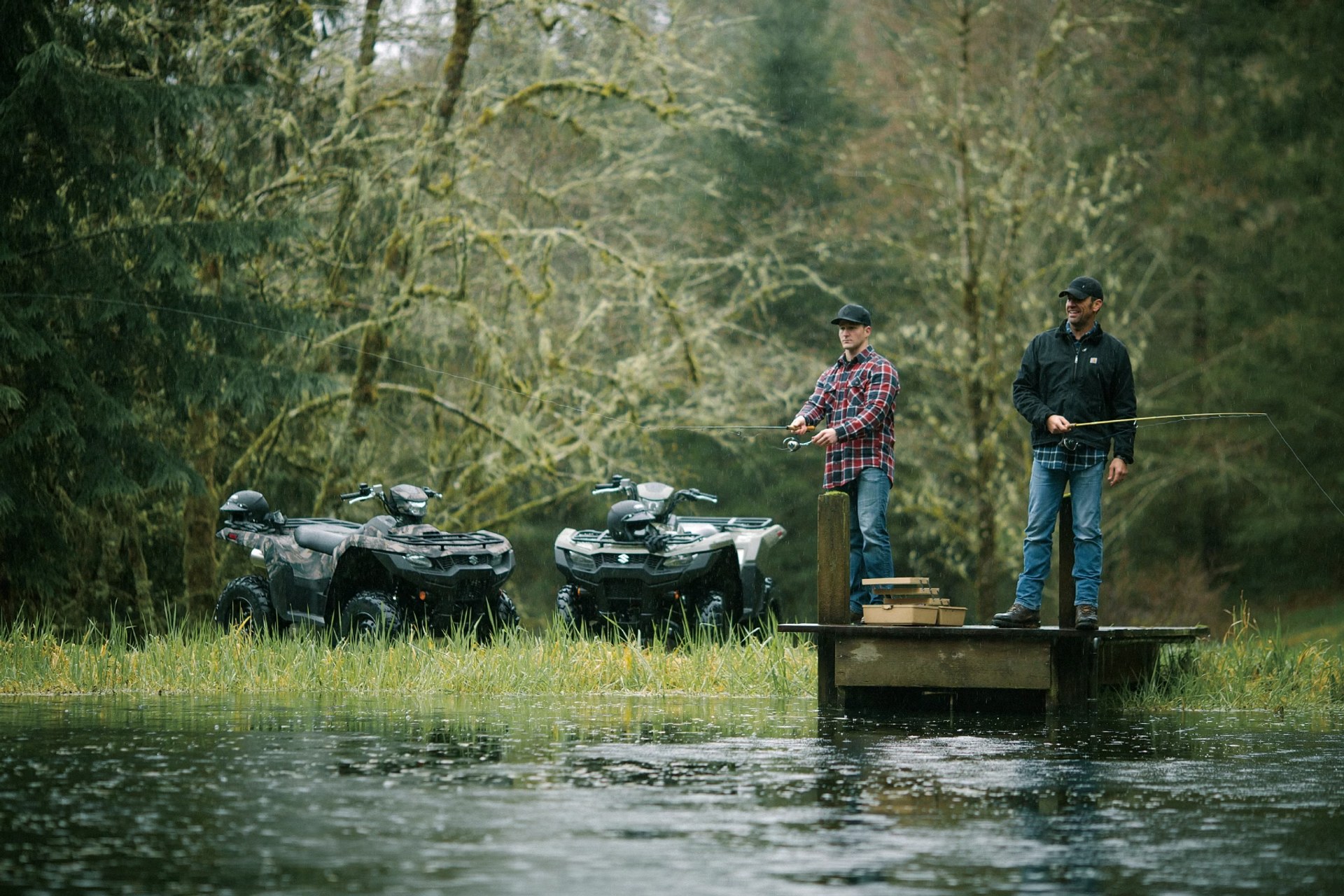 Dos personas pescando junto a cuatrimotos Suzuki KingQuad 750 en un lago rodeado de bosque