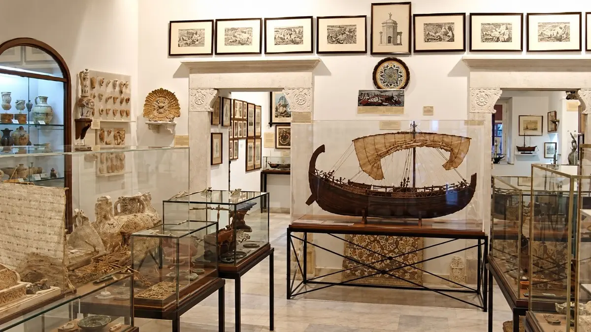 Interior of the Aegean Maritime Museum in Mykonos, featuring a detailed model sailing ship, glass display cases, historic artefacts, ceramics and framed maritime artwork.