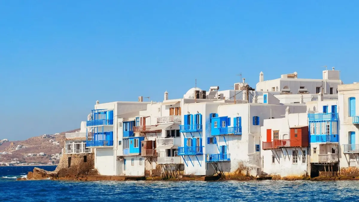 Little Venice in Mykonos with whitewashed waterfront houses, colourful blue and red balconies, and the Aegean Sea under a clear blue sky.